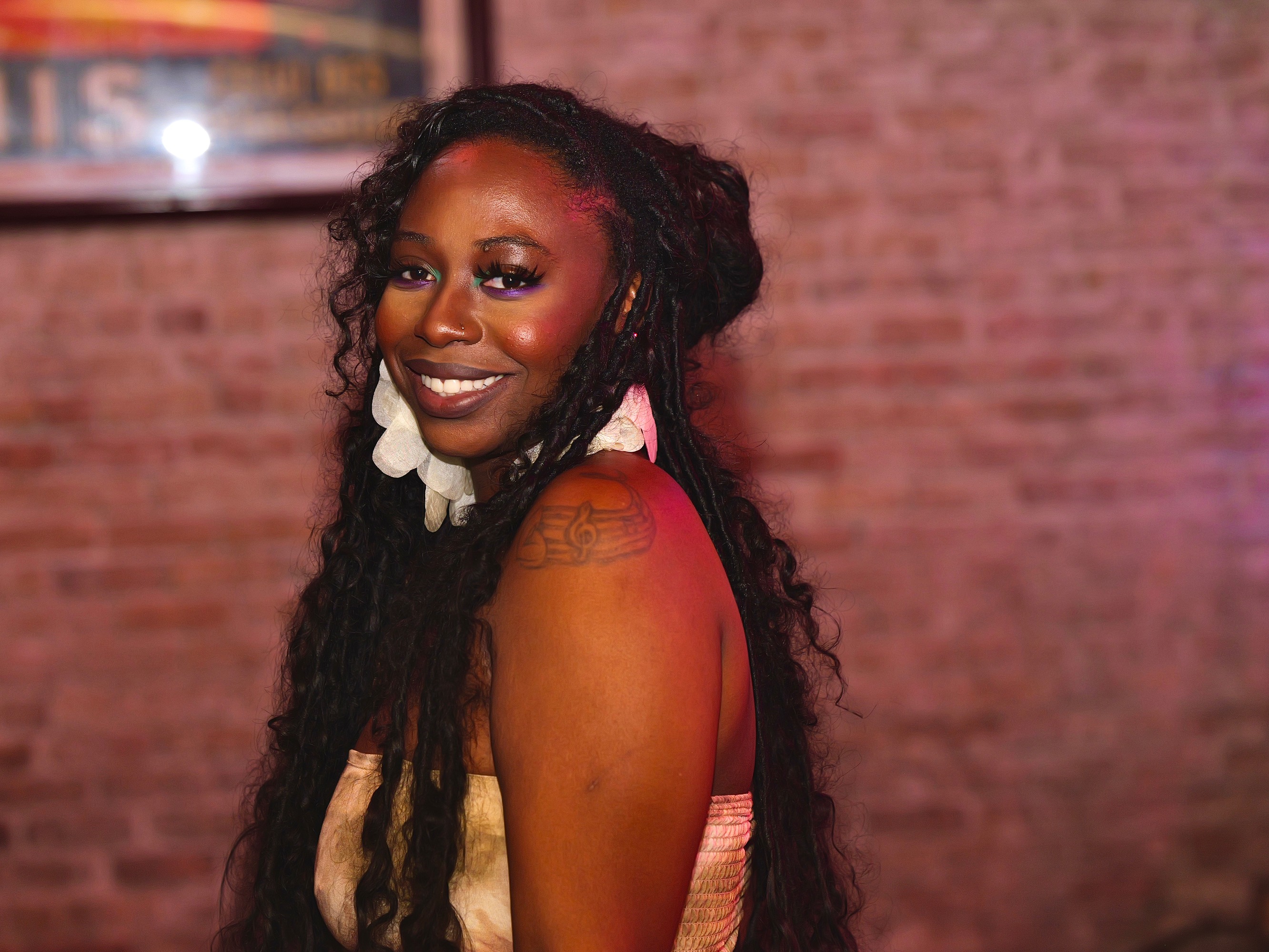 DVINE smiling against a brick wall, music note tattoo visible on her shoulder