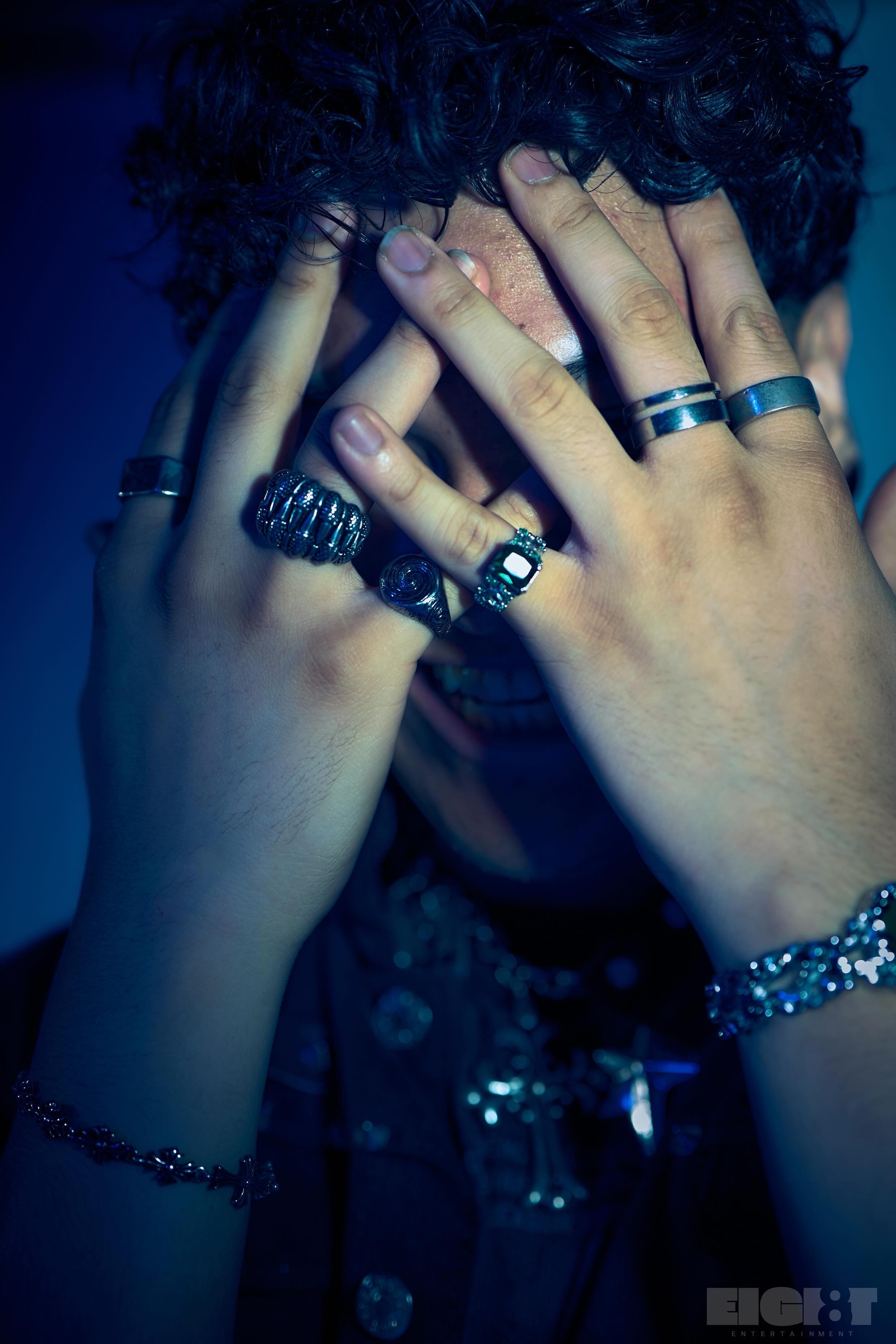 Evver the Thinker in a moody blue-lit shot, hands raised to his face, wearing multiple rings