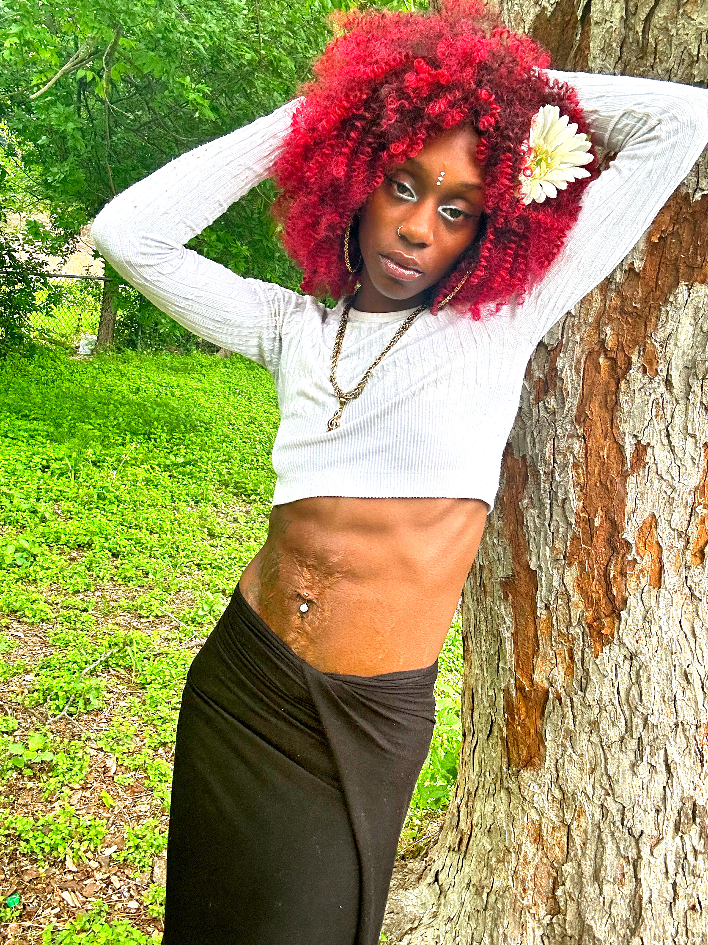 Ife Devine portrait, red curly hair with white flower, leaning against a tree