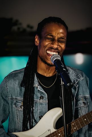 Yadin Kol at RRL 2026, with long locs in denim jacket smiling at the mic with white guitar in teal glow