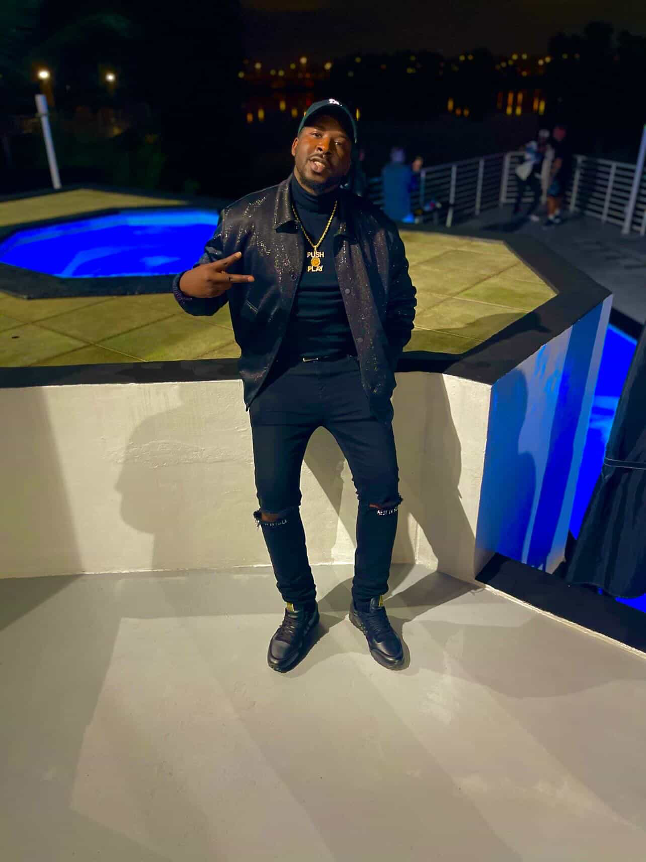 PUSHPLAY914 in a black sequined jacket and gold Push Play chain, posed at a rooftop pool at night