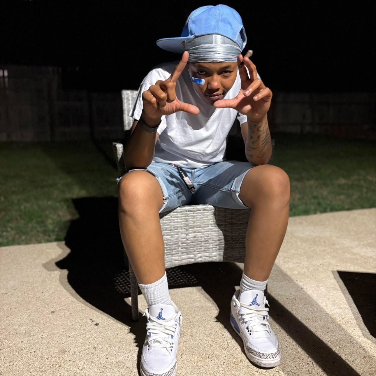 TGN Lank$ seated outdoors in a blue cap, throwing up hand signs